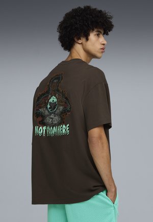 Junger Mann mit lockigem Haar trägt ein braunes, übergroßes T-Shirt mit einer Grafik einer Figur und dem Text „NOT FROM HERE“ auf dem Rücken, mintfarbene Shorts.