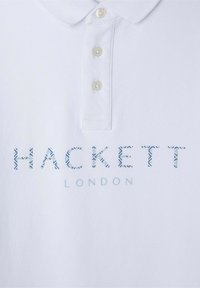 Biela polokošeľa s textúrovanou tkaninou. Má tri gombíky na zapínanie a modro potlačené logo s vzorom, ktoré znie "HACKETT LONDON."