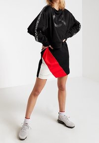 Schwarzes Sweatshirt mit langen Ärmeln und Logo auf der Brust, kombiniert mit einem knielangen Rock, der diagonale Farbschattierungen in Rot, Schwarz und Weiß aufweist.