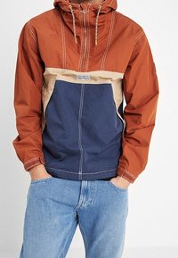 Windbreakerjas in roestoranje, beige en donkerblauw. Heeft een halve rits aan de voorkant, een capuchon en contrasterende stiksels in een losse pasvorm.