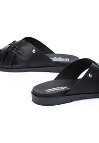 Sandalias deslizantes de cuero negro con diseño de tiras cruzadas, que presentan acentos en tono dorado y una suela texturizada negra. Planas y con punta abierta.