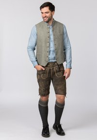 Die traditionelle Kleidung für Männer umfasst ein hellblaues Hemd mit Knöpfen, eine olivgrüne Weste, braune Shorts mit Stickereien und kniehohe dunkle Socken.