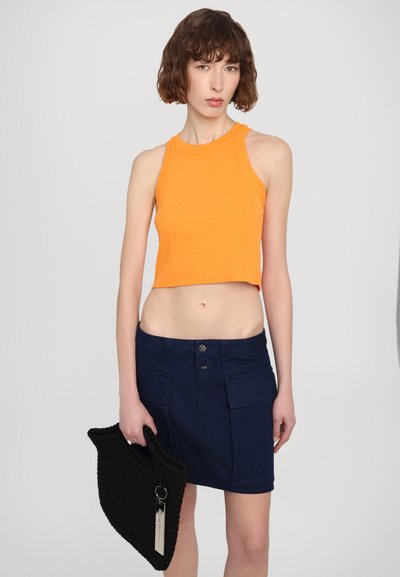Oranžs ribots īsais tops, kas saskaņots ar tumši zilu džinsu svārku ar priekšējiem kabatām. Modelis tur melnu aušanu somu. Vienkārša fona.