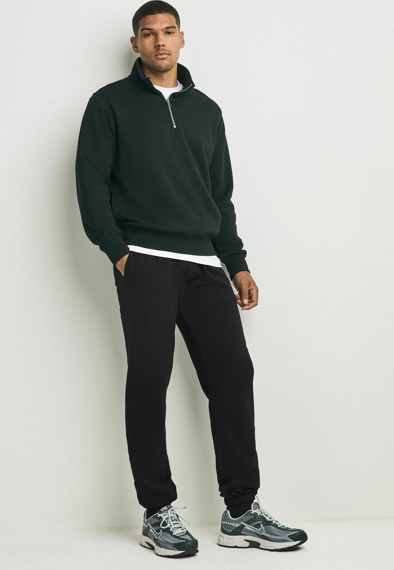 Schwarze Zip-Up-Sweatjacke mit gerippten Bündchen, kombiniert mit schwarzen Jogginghosen und grauen Sportschuhen. Glattes Material und lässige Passform.