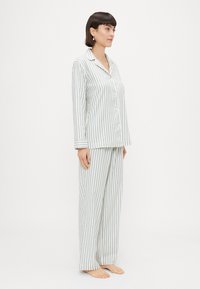 Pajama set s prugicama u laganom materijalu. Košulja na dugme s dugim rukavima, ovratnikom i odgovarajućim hlačama. Boje: bijela s zelenim i plavim prugama.