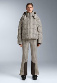 Žena stojaca v puffer kabáte s houndstooth vzorom a zodpovedajúcich flared nohaviciach s čiernymi látkovými panelmi na dolných nohách, obutá v čiernych topánkach.