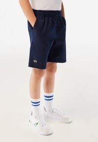 Pantaloni corti blu navy con vita elastica, tasche laterali e logo ricamato verde. Abbinati a calzettoni bianchi con righe blu e scarpe bianche.