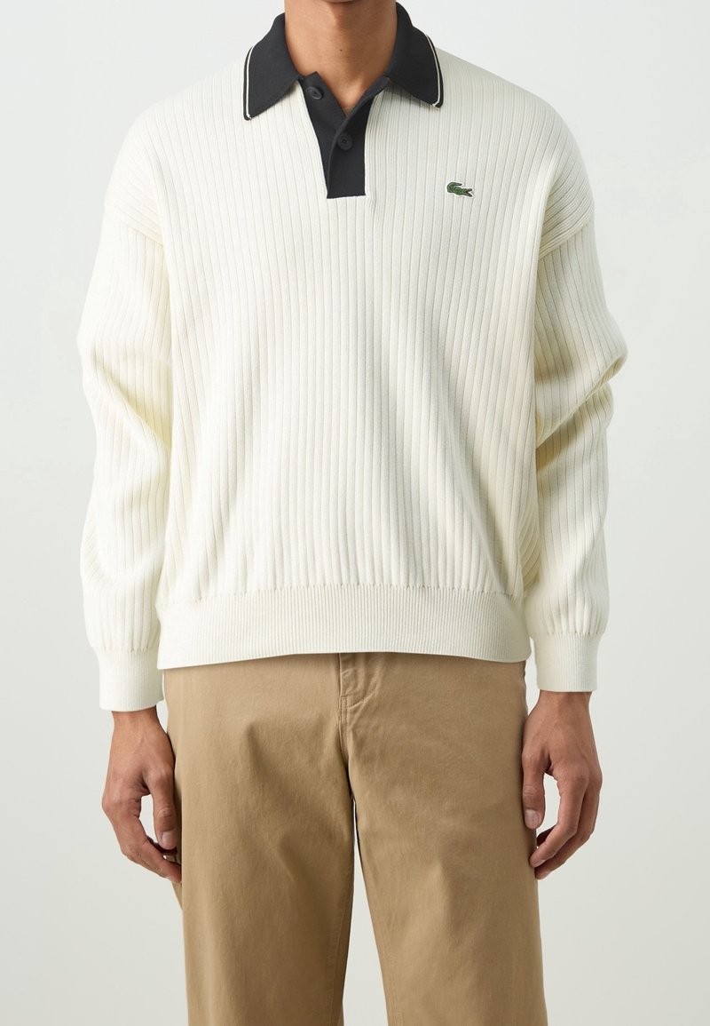 Homme portant un pull côtelé blanc avec un col polo noir et un pantalon beige, debout devant un fond clair uni.