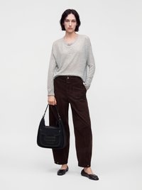 Femme aux cheveux courts et foncés portant un haut gris clair à manches longues, un pantalon large sombre, des ballerines noires, tenant un sac à épaule noir.