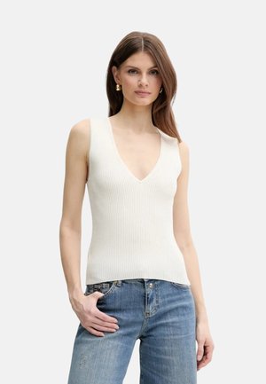 Donna che indossa un top bianco senza maniche a coste con scollo a V e jeans blu, posando con una mano in tasca su uno sfondo uniforme.