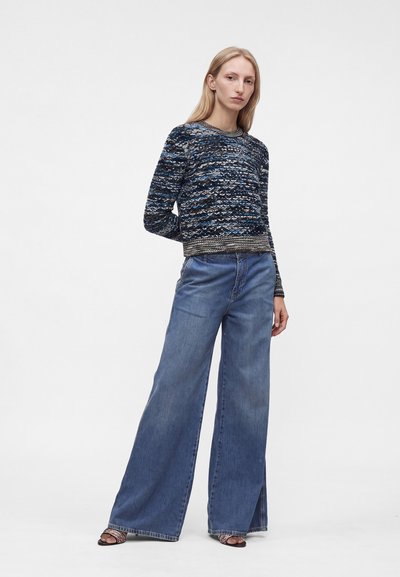 Femme portant un pull sombre à motifs, un jean bleu à jambes larges et des talons ouverts, debout avec une main dans le dos sur un fond blanc uni.