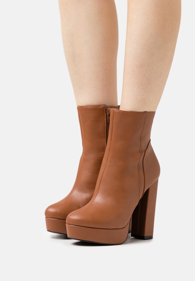 Jambes portant des bottes plateformes en cuir brun mi-mollet avec des talons hauts épais et des fermetures éclairs latérales sur un fond uni.
