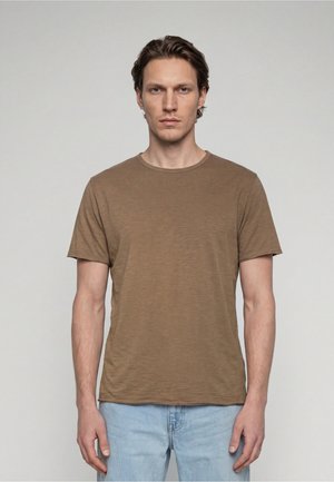 Giovane uomo con capelli castani corti che indossa una maglietta marrone tinta unita a maniche corte e jeans azzurri, in piedi davanti a uno sfondo grigio uniforme.
