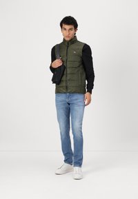 Maslinasto zeleni puffer prsluk s visokom kragnom, crne dugih rukava, svijetloplave traperice i bijele tenisice. Mali crni crossbody ranac.