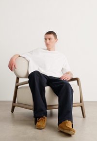 White t-shirt with subtle branding, dark blue pants, seated on a light fabric chair with wooden frame. Neutral background, minimalist design.