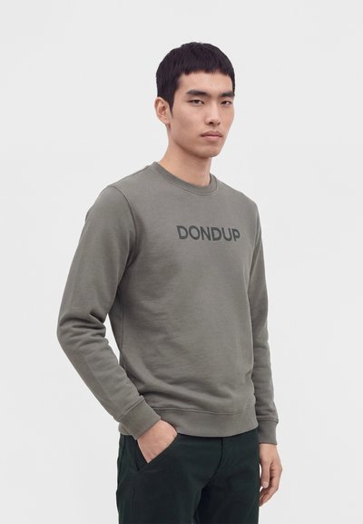 Homme portant un sweat-shirt vert olive avec « "DONDUP" » imprimé sur la poitrine et un pantalon foncé, debout avec une main dans la poche.