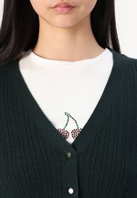 Dark green knitted cardigan with a V-neck, featuring subtle texture and gold-colored buttons, over a white T-shirt with cherry embellishments.