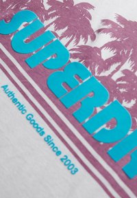 Camiseta blanca con el texto en negrita color turquesa "SUPERDRY", un diseño de palmera morada y el texto "Productos Auténticos Desde 2003".