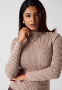 Maglione beige a coste con collo alto, caratterizzato da dettagli in metallo dorato sulla spalla e motivo a maglia strutturato su tutta la superficie.