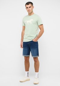 Miętowy t-shirt z białym nadrukiem konia, w połączeniu z niebieskimi szortami dżinsowymi. Białe trampki i wzorzyste skarpetki dopełniają strój.