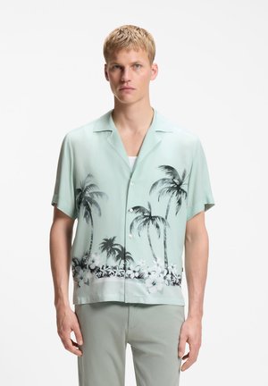 Ung mand iført en lysegrøn kortærmet skjorte med sort palme- og hvid blomstermønster, kombineret med lyse bukser.