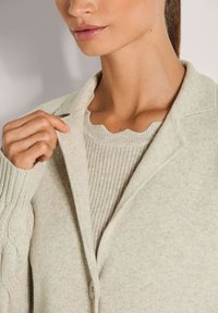 Frau, die eine hellbeige Strickjacke über einem gerippten Pullover mit gewelltem Halsausschnitt anpasst, gezeigt von Hals bis Brust.