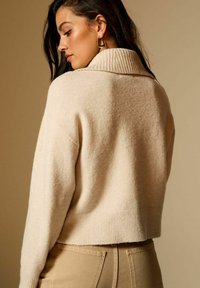 Suéter de punto beige con cuello acanalado, diseño corto y tejido texturizado. La espalda del modelo se muestra contra un fondo de tonos cálidos.