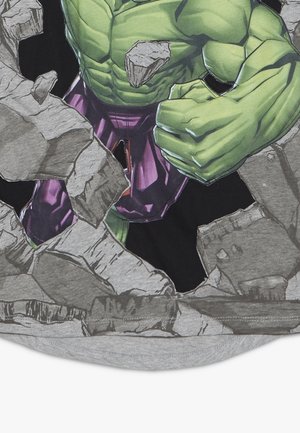 T-shirt gris mettant en scène une figure humanoïde verte traversant une surface rocheuse. La figure porte un pantalon violet avec des textures détaillées et des ombrages.