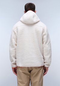 Man som bär en beige fleecejacka med huva, vänd bort från kameran mot en enkel ljusgrå bakgrund.