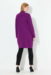 Langer lila Cardigan mit flachem Kragen und langen Ärmeln, kombiniert mit schwarzen Leggings und schwarzen High Heels. Glatte Textur.