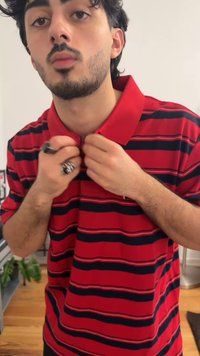Young man adjusting collar of red and black striped polo shirt, wearing rings on fingers, standing indoors with plant and light background.