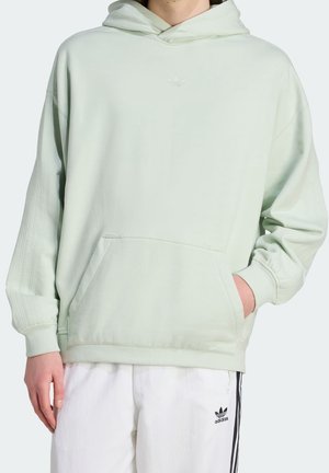 Osoba ubrana w jasnobieżący zielony kaptur Adidas z kieszenią kangurką oraz białe spodnie dresowe Adidas z czarnymi paskami.