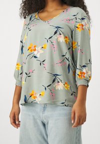 Donna che indossa una blusa floreale a maniche lunghe di un verde chiaro con polsini arricciati e jeans azzurri, in piedi con le braccia rilassate.