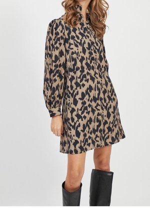 Femme portant une robe à manches longues imprimée animal beige et noir, associée à des bottes noires montantes jusqu'aux genoux, sur un fond uni.