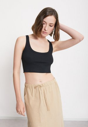 Jeune femme aux cheveux courts bruns portant un crop top noir et une jupe beige, posant avec un bras levé derrière la tête.