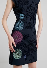 Granatowa sukienka z misternym haftem kwiatowym w wielokolorowych wzorach. Posiada rękawy kimono i dopasowany krój. Gładka tekstura materiału.