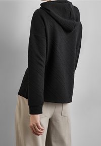 Sweat à capuche noir avec un motif en diamants texturé, manches longues et coupe ample, associé à un pantalon beige clair.