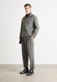 Grau karierter Blazer mit zwei aufgesetzten Taschen, kombiniert mit lockeren grau karierten Hosen. Schwarze Schuhe runden den Look ab.