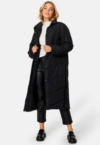 Lang sort dunjakke med knaplukning fortil, høj krave og quiltet tekstur. Båret over en lys sweater og sorte læderbukser.