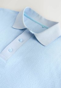 Polo bleu clair avec un motif texturé en zigzag et lignes, plié à plat, montrant le col et deux boutons.