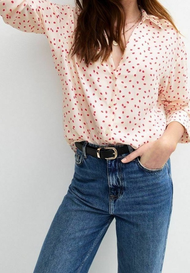 Blouse crème à pois rouges, manches longues et col, rentrée dans un jean en denim bleu taille haute avec une ceinture noire.