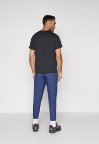 Άνδρας που στέκεται με την πλάτη προς το κοινό, φορώντας ένα μαύρο μπλουζάκι κοντομάνικο, μπλε παντελόνια, λευκές κάλτσες και μαύρα αθλητικά παπούτσια, με φόντο μια απλή επιφάνεια.