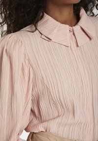 Blusa texturizzata rosa chiaro con colletto classico, maniche arricciate e sottili pieghe verticali. Include un dettaglio di un singolo bottone decorativo.