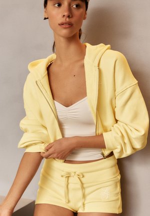 Ung kvinde justerer ærmerne på en gul zip-up hættetrøje, iført matchende gule shorts og en hvid rynket cropped top mod en neutral baggrund.