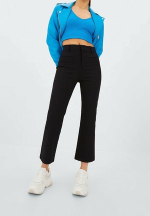 Vrouw die een felblauwe cropped jas en top draagt met hoogtalige zwarte broek en witte chunky sneakers, staand tegen een effen achtergrond.