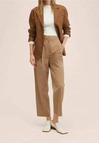 Blazer marron sur un pull léger, associé à un pantalon beige. Des mocassins blancs complètent la tenue. Le matériau semble texturé, avec une coupe ajustée.