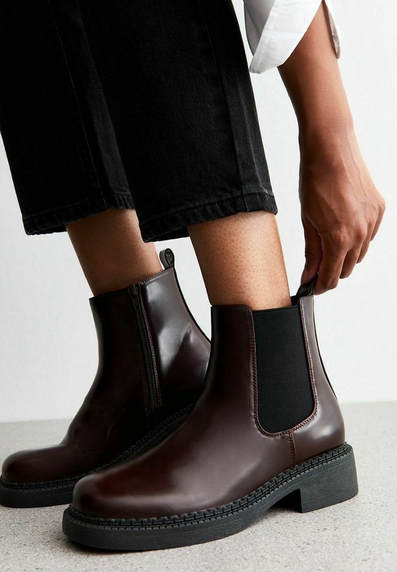 Personne portant des bottines Chelsea marron brillantes et un pantalon noir raccourci, tirant sur la languette de la botte en se tenant sur un sol clair.