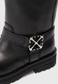 Botín de cuero negro con suela resistente y detalle decorativo de hebilla de metal plateado en la correa del tobillo, vista de cerca.