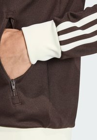 Hnedá športová bunda s bielymi pruhovanými akcentmi na rukávoch, štruktúrovanou látkou, zipsovou kapsou a priliehavými bielymi manžetami.