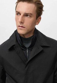Abrigo negro con un cuello afilado, hecho de una tela suave. Cuenta con una capa interna con cremallera y una textura sutil en el cuello y los puños.